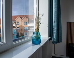 Una ventana con un jarrón azul que contiene plantas secas, vista de un edificio con balcones y un radiador en la pared.