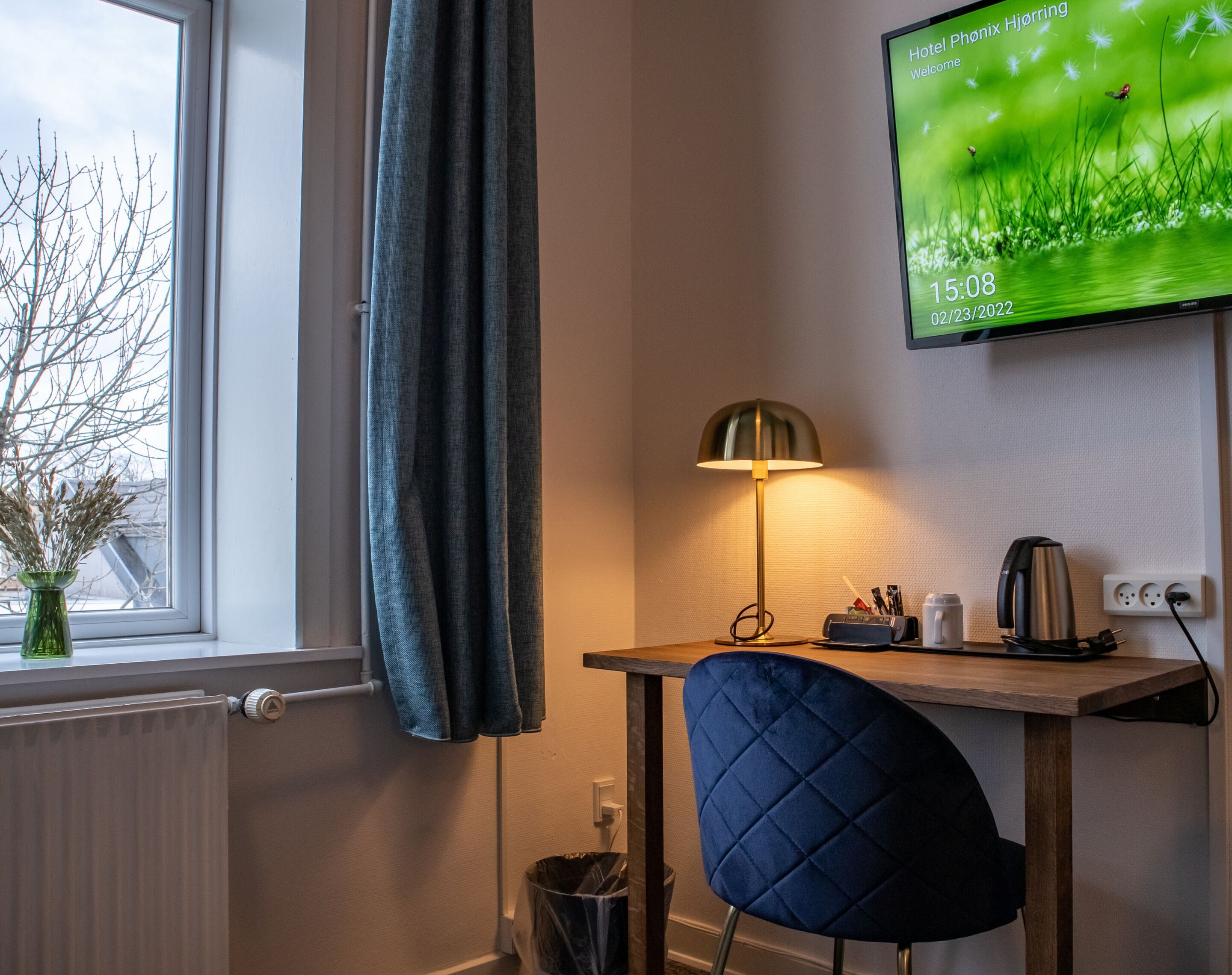 Habitación de hotel con escritorio, una silla, una lámpara, un televisor mostrando un mensaje de bienvenida y una ventana con vista a árboles desnudos.