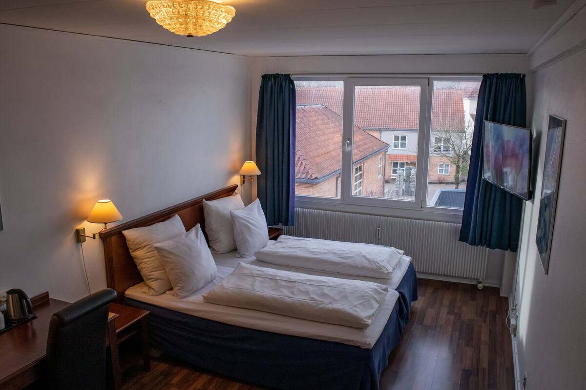 A hotel room with a double bed, two bedside lamps, a desk with a chair, a radiator, and a window with curtains.