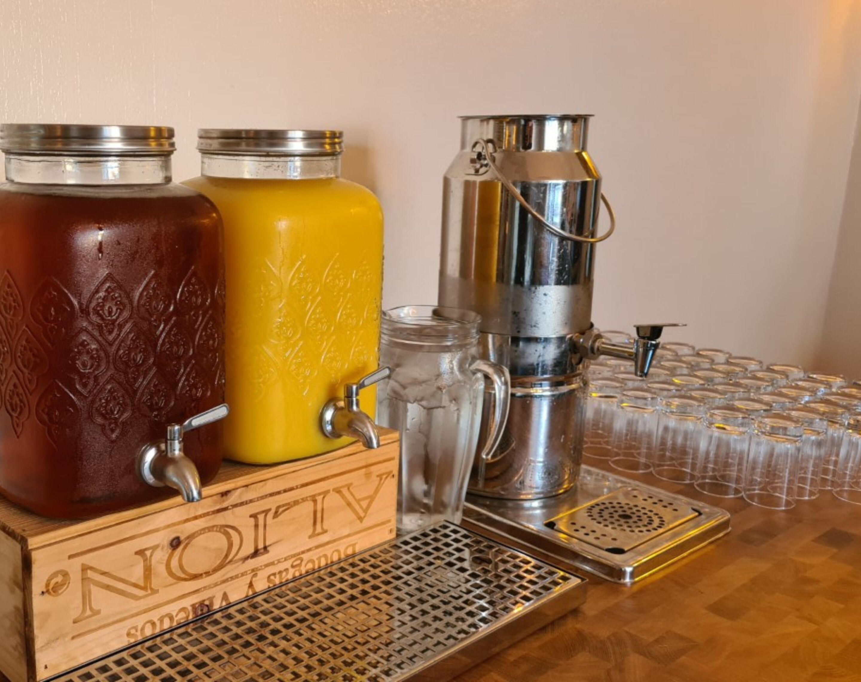 Una barra equipada con tres dispensadores de bebidas, una jarra de vidrio y una bandeja con vasos vacíos.