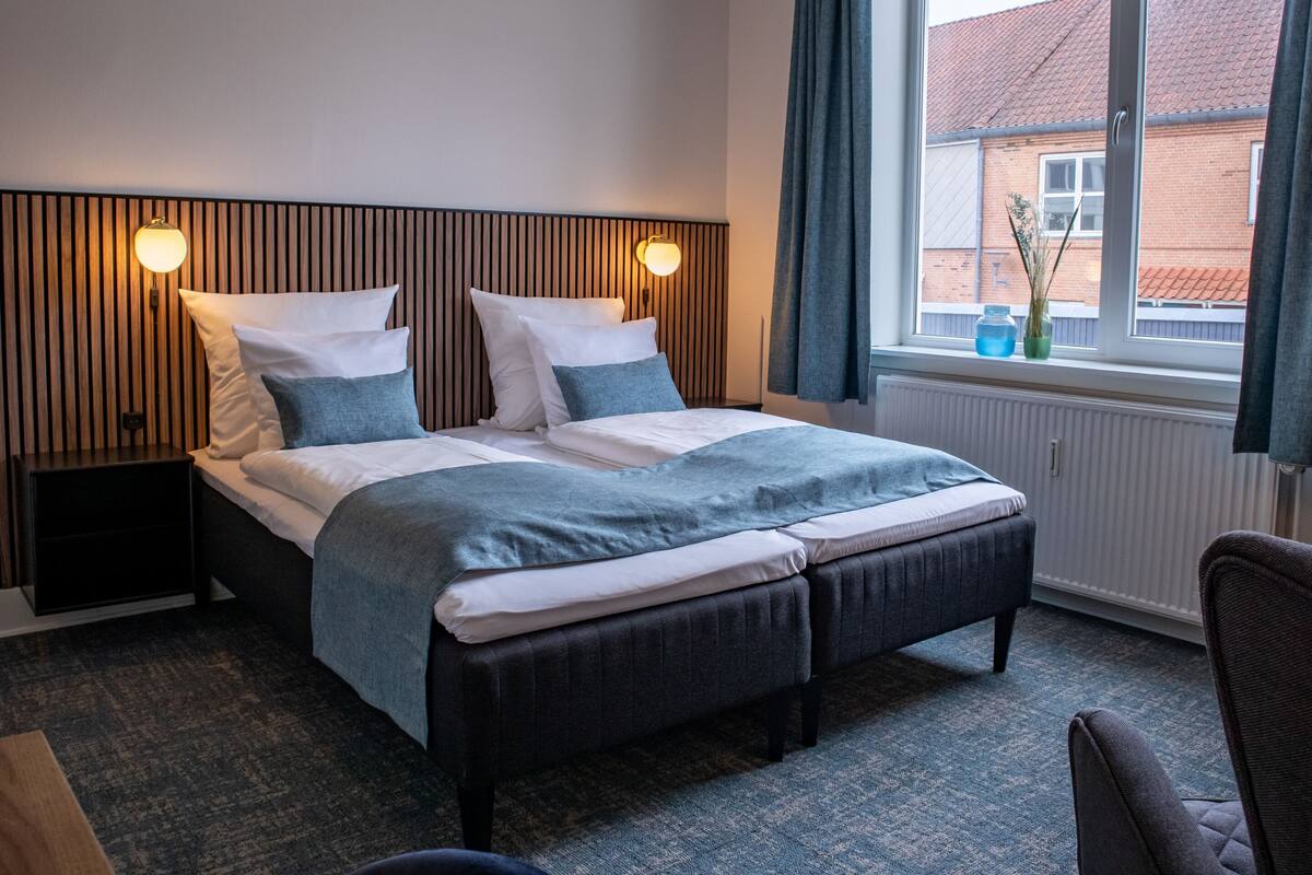 A hotel room with a bed, bedside tables, a window with curtains, and a view of a building outside.