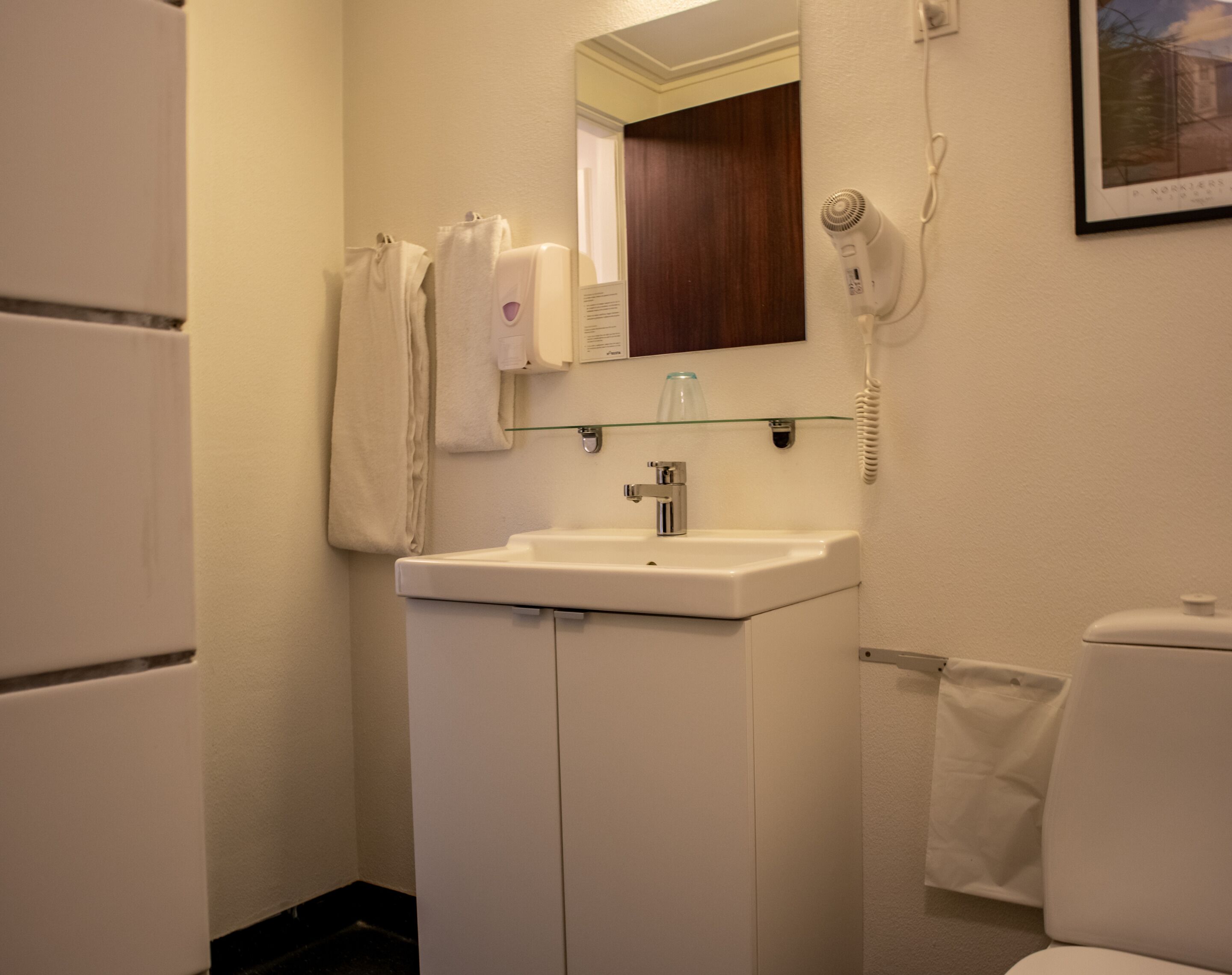 A small, clean bathroom with a white sink, a mirror above it, a hairdryer on the wall, and a toilet.