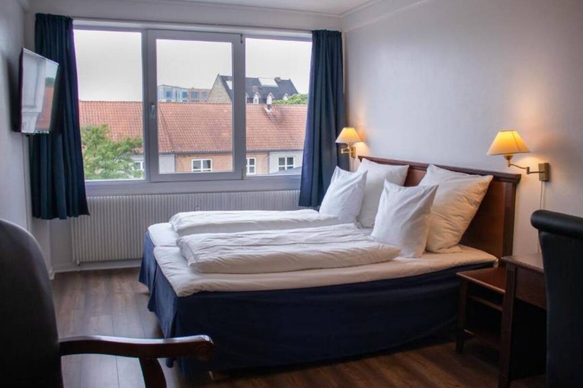 A hotel room with two beds, a TV, a window with curtains, and a lamp on the nightstand.