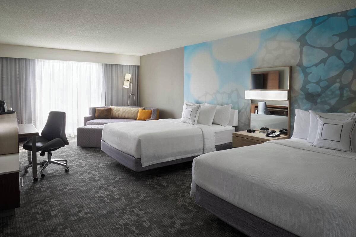 A hotel room with two beds, a desk, a chair, and a large window with curtains.