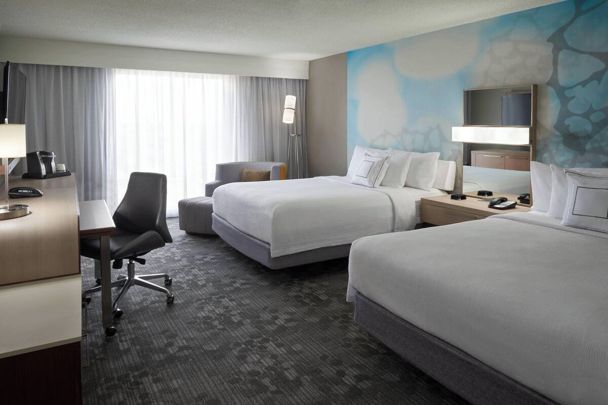 A hotel room with two beds, a desk, a chair, a lamp, and a TV.
