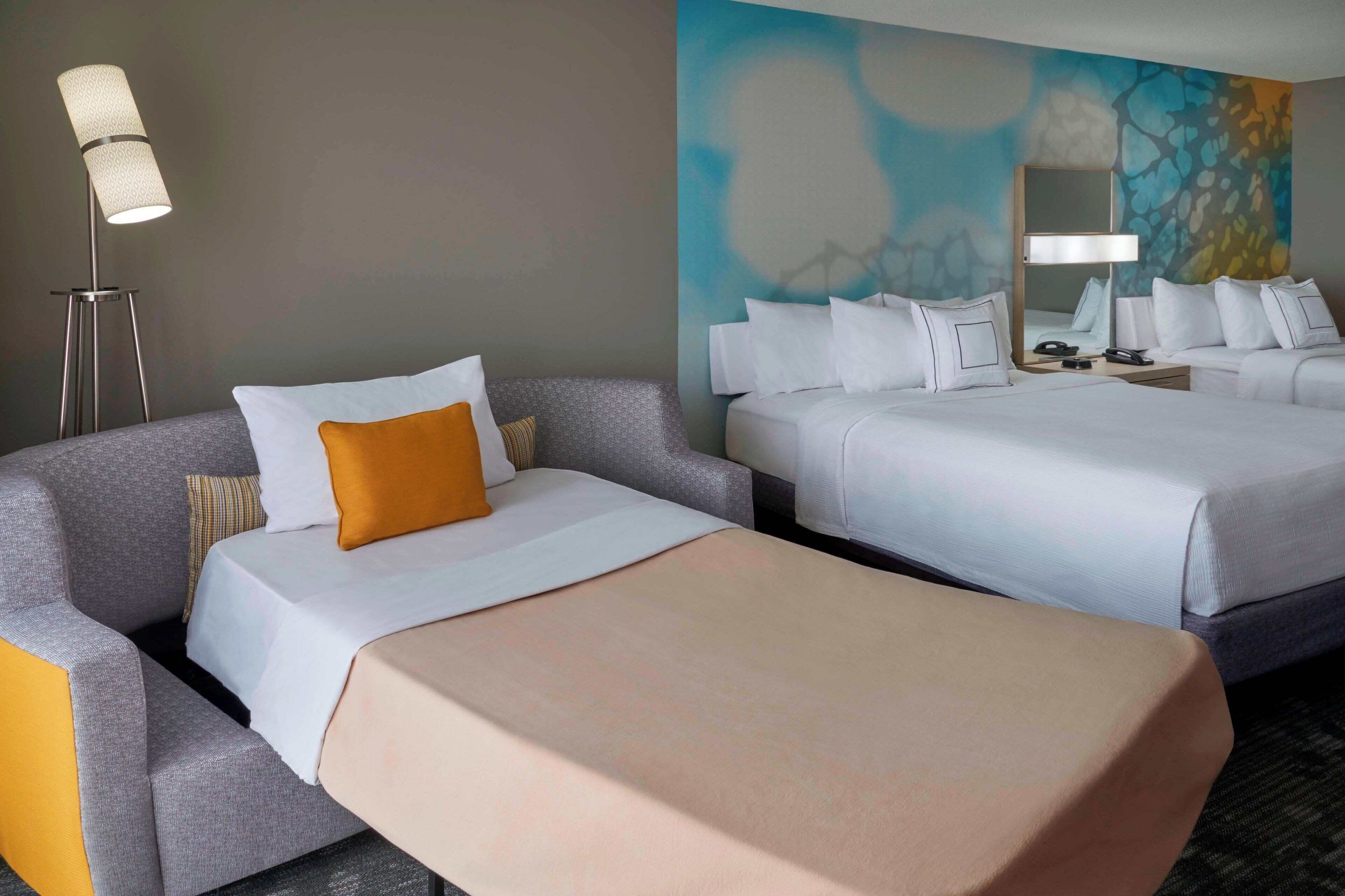 Habitación de hotel con dos camas, un sofá, una lámpara de pie y un cuadro abstracto grande en la pared.