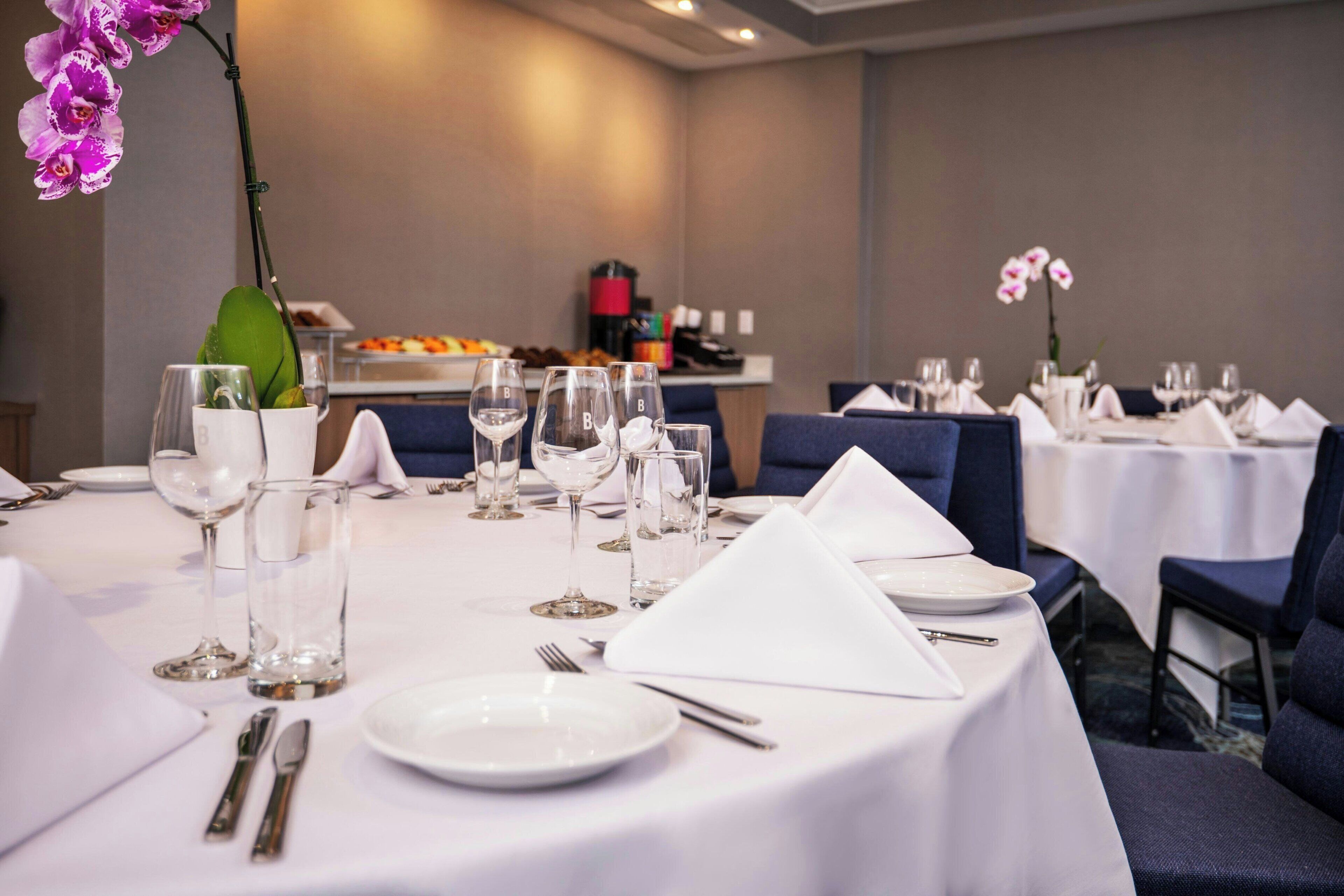 Une table à manger dressée avec des nappes blanches, des assiettes, des verres et des couverts, et un vase d’orchidées placé sur le côté gauche.