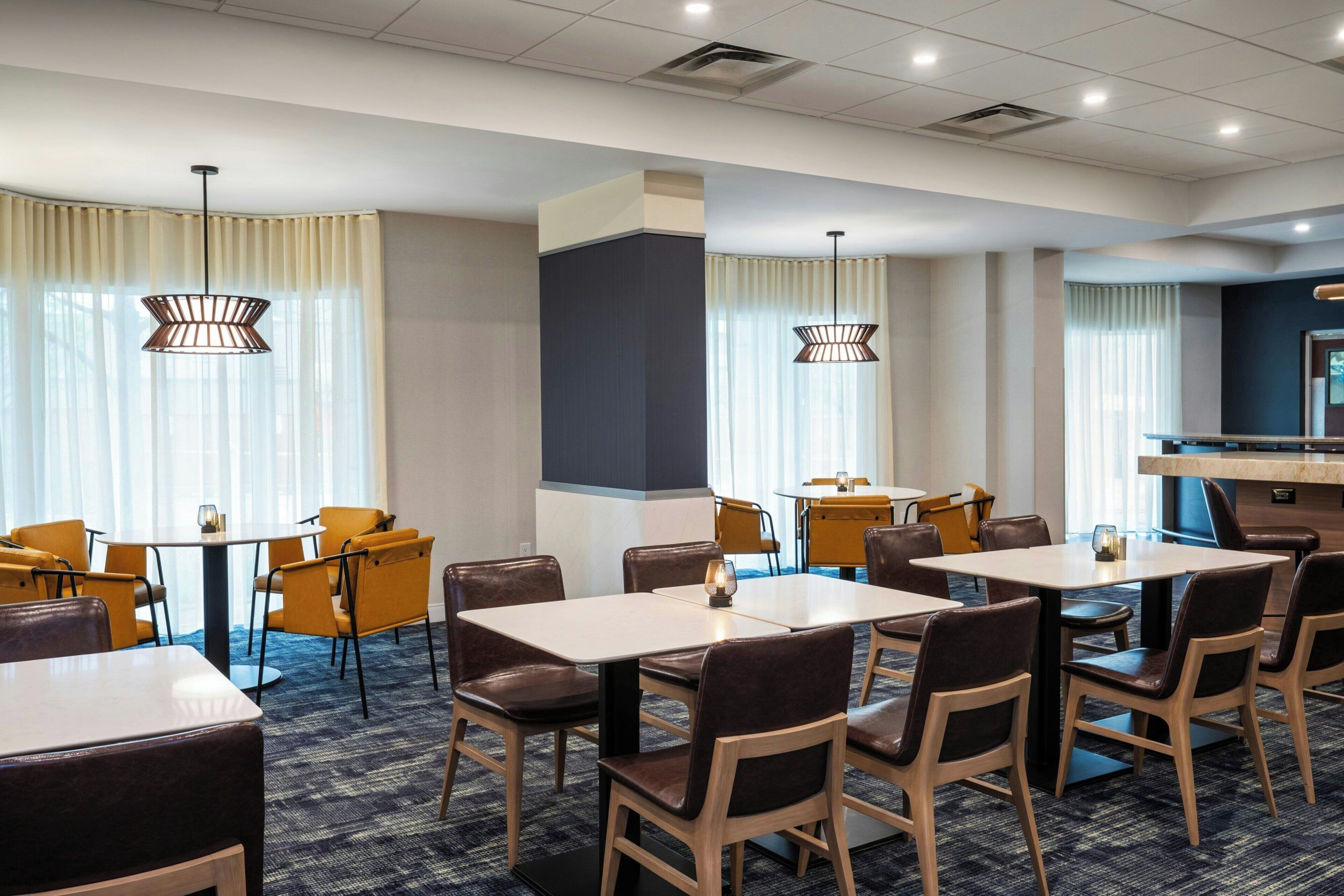 A hotel dining area with multiple tables and chairs, large windows with curtains, and pendant lighting.