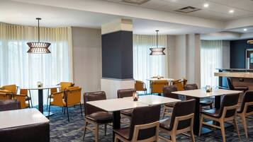 A hotel dining area with multiple tables and chairs, large windows with curtains, and pendant lighting.