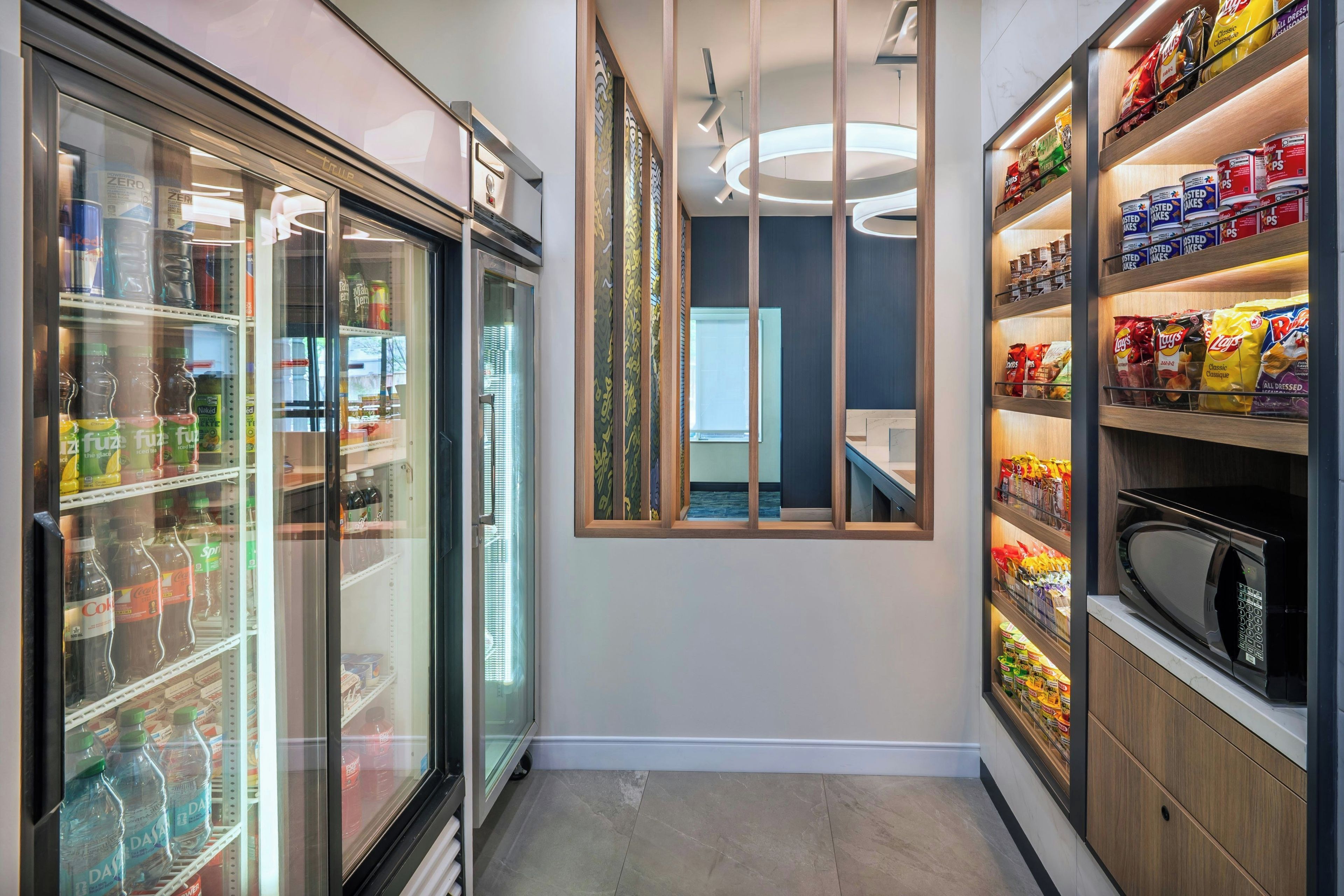 Un pasillo con un refrigerador de bebidas, un microondas y estantes con aperitivos.