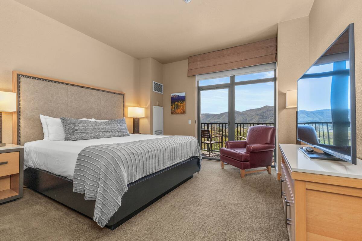 A hotel room with a large bed, a TV, a desk, a chair, and a balcony with a view of the mountains.