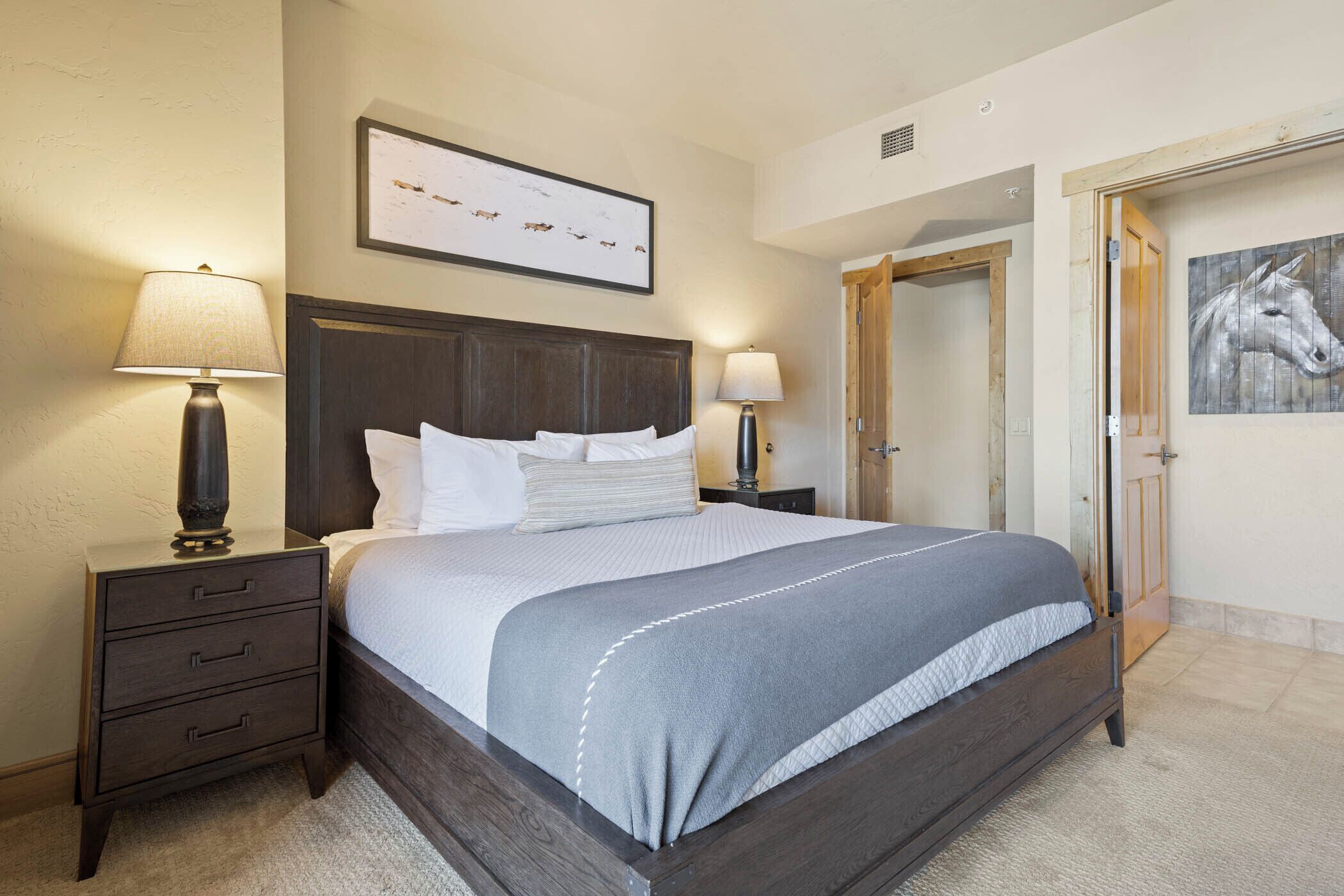 A neatly made bed with a dark headboard, two bedside tables with lamps, a framed picture above the bed, and a wooden door to the right.