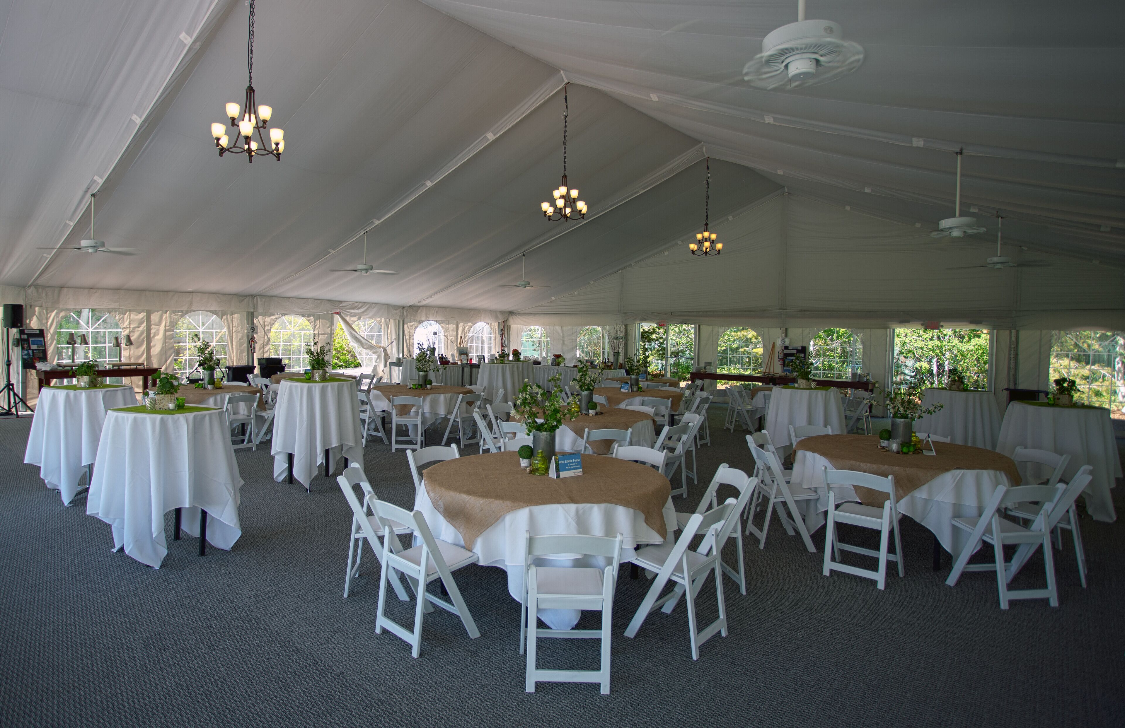 Un amplio espacio para eventos con carpas, con varias mesas redondas cubiertas con manteles blancos y rodeadas de sillas blancas.