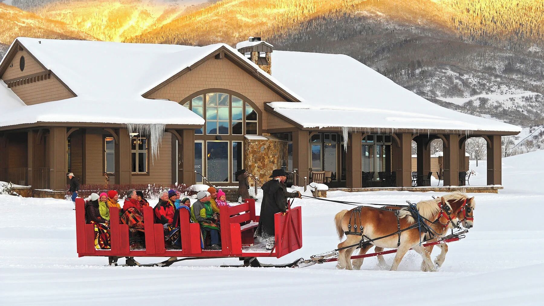 A lodge with a snow-covered roof, large windows, and a stone chimney.