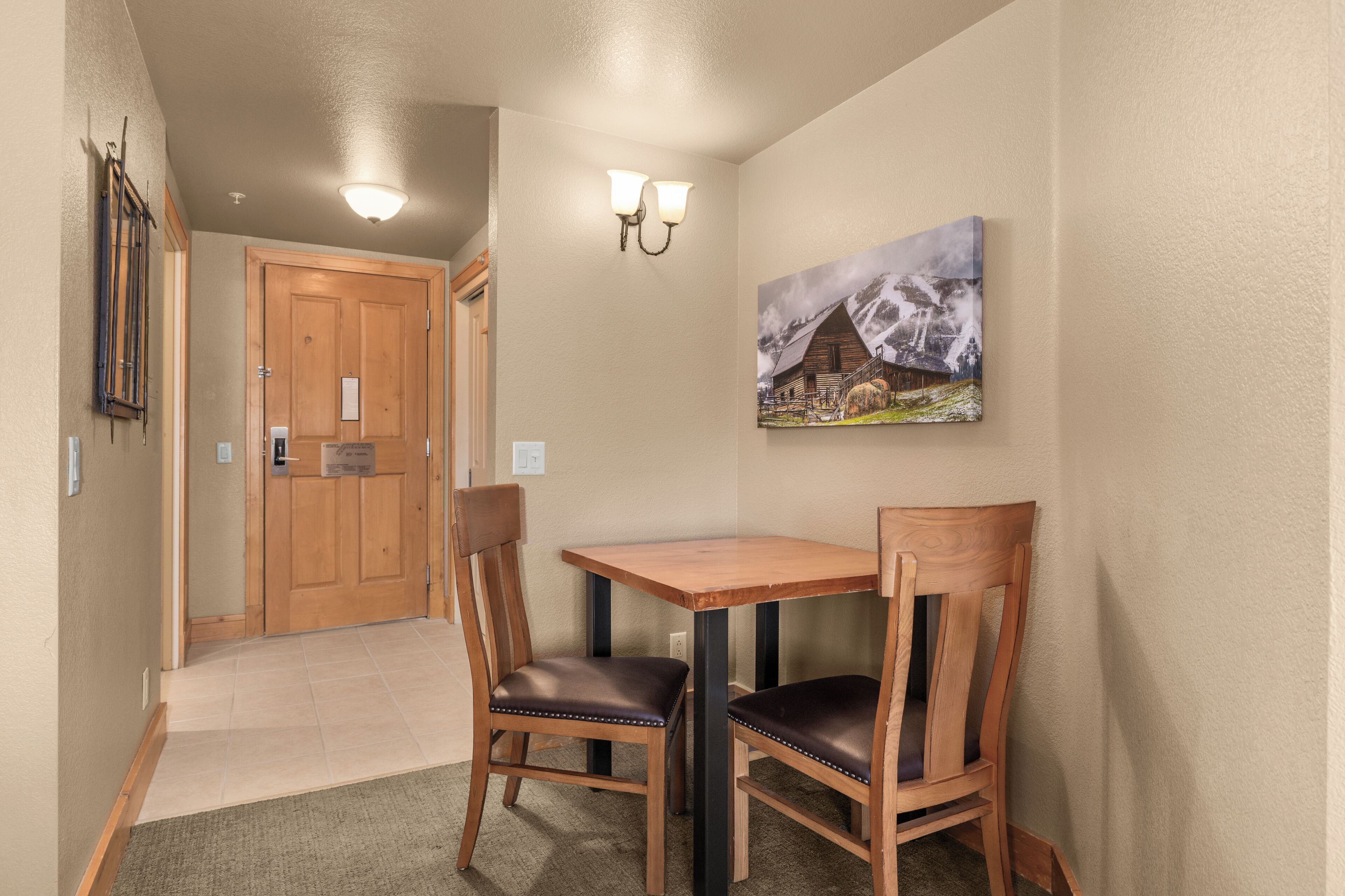 Un couloir avec une porte en bois, une petite table avec deux chaises et un tableau accroché au mur.