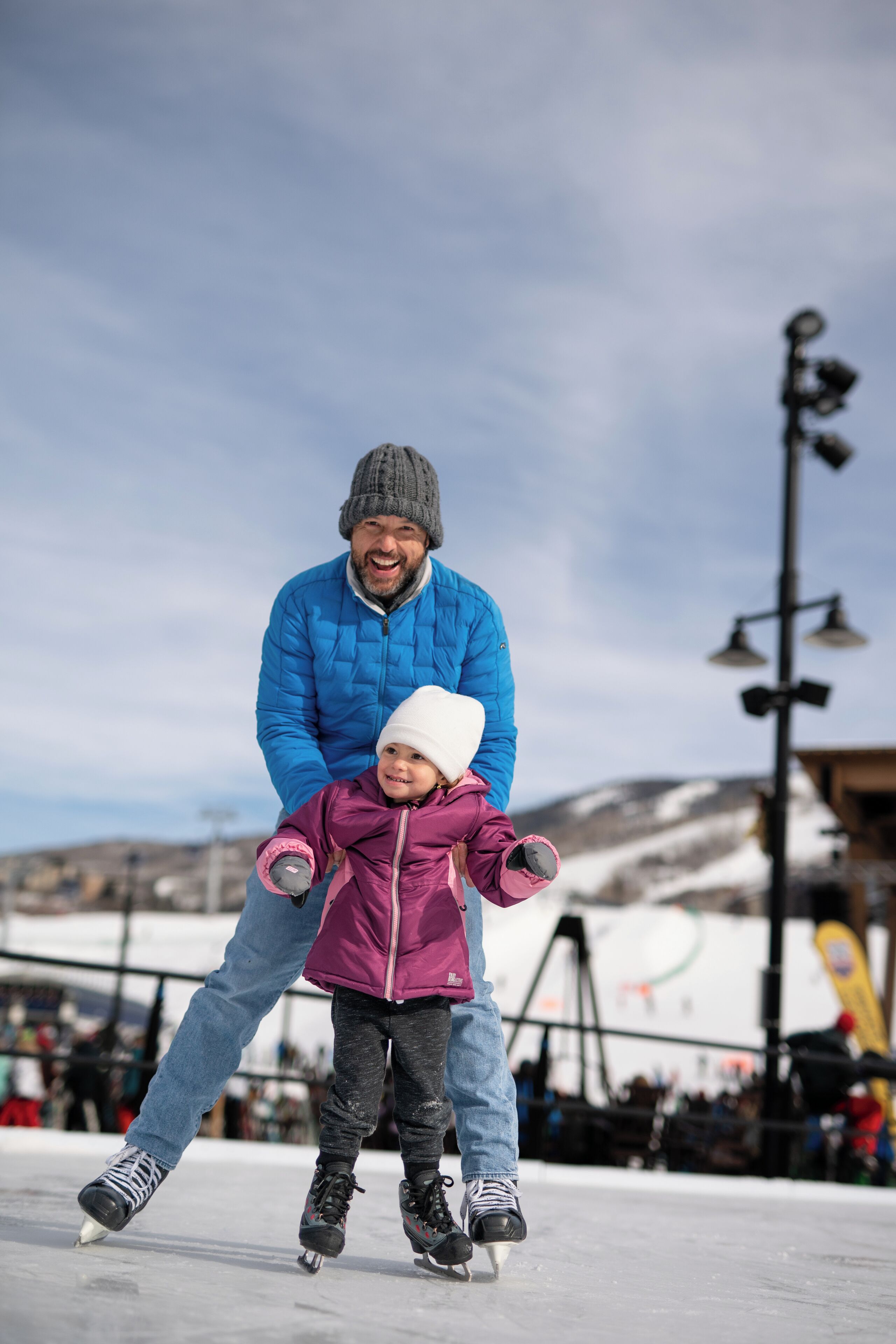 Una persona con chaqueta azul y gorro gris está detrás de un niño con chaqueta morada y gorro blanco, ambos sobre patines.