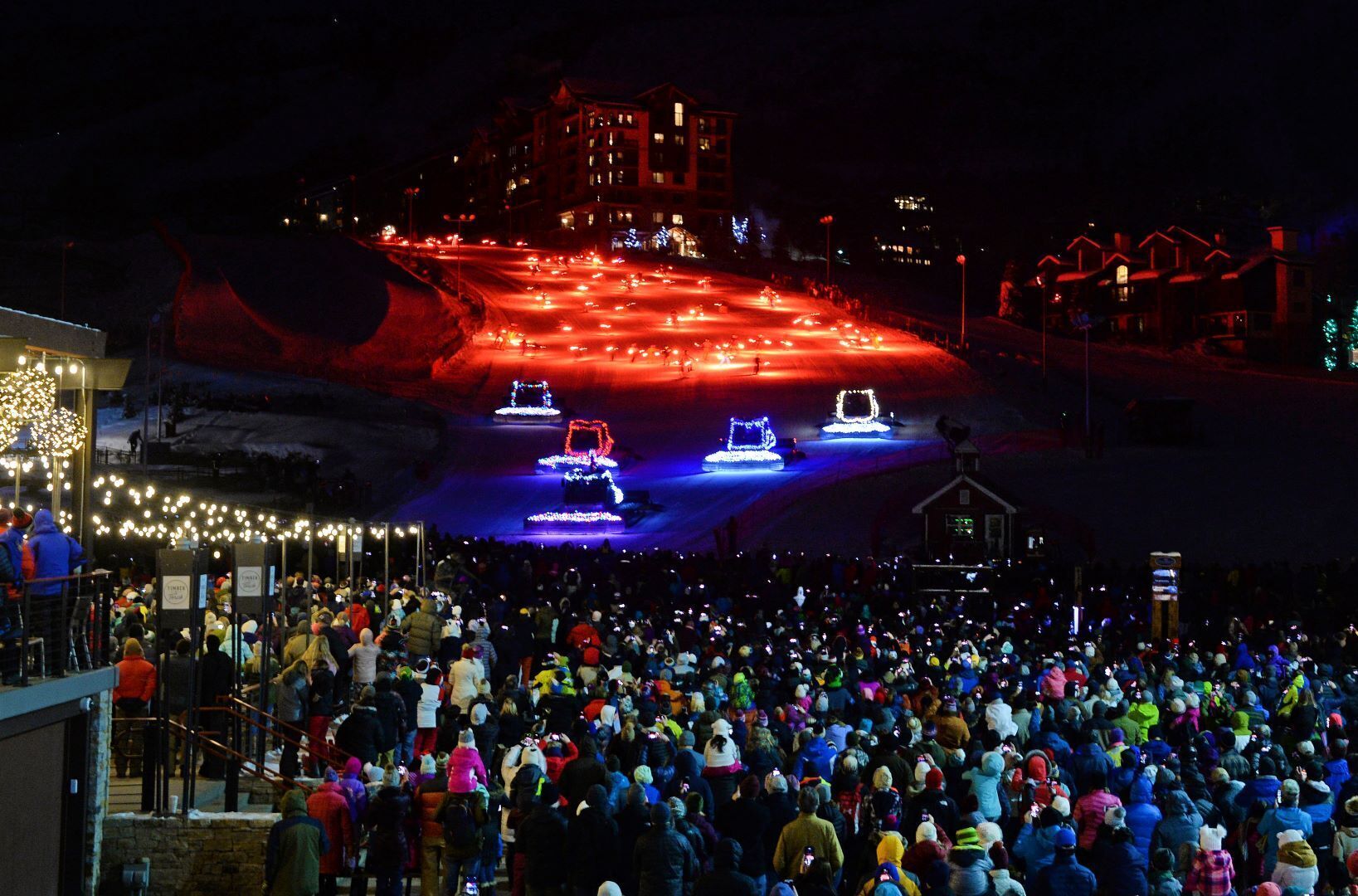 A large crowd gathered at a ski resort with illuminated ski lifts and buildings in the background.