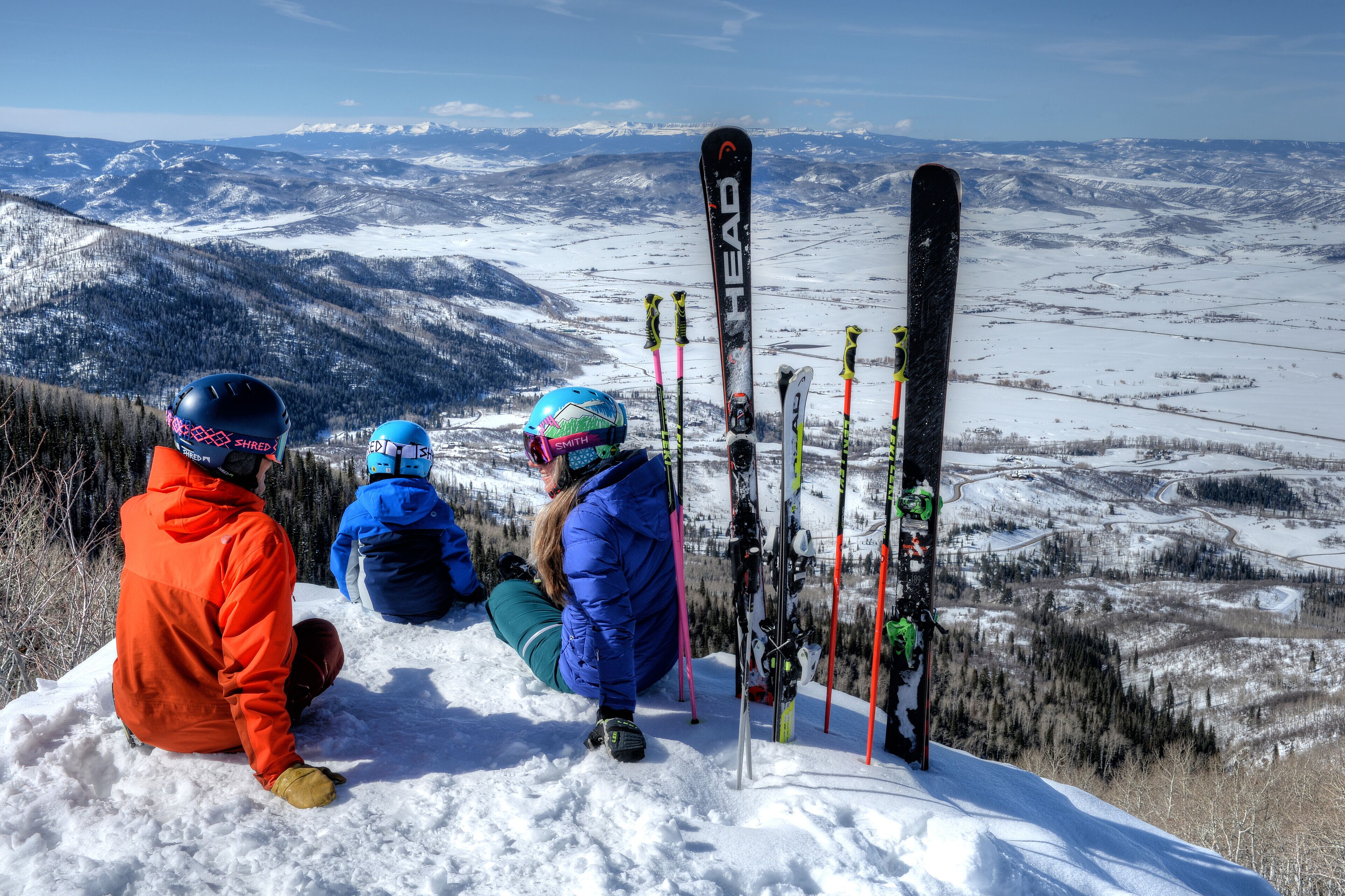 Three people sitting on a snowy slope with skis leaning against a snow-covered hill.