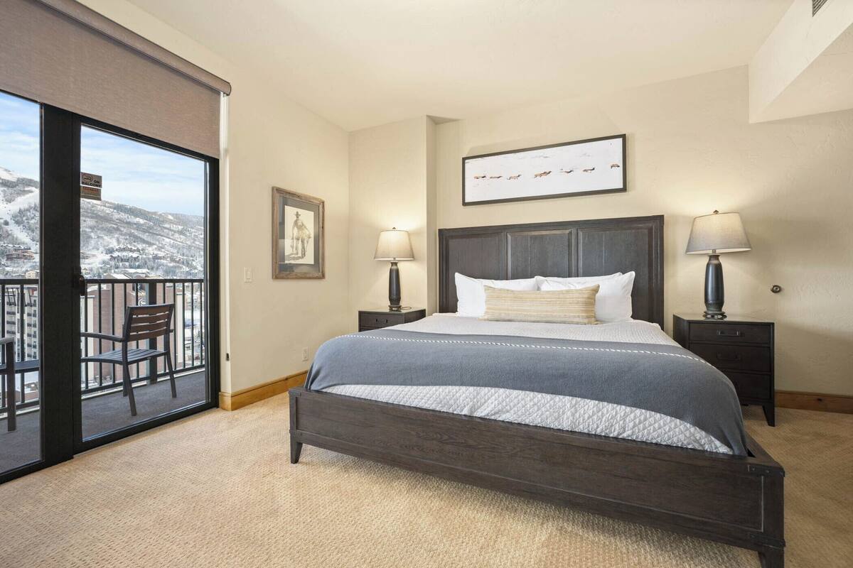 A bedroom with a large bed, two bedside tables, a chair, a balcony with a view of a snowy landscape, and a framed picture on the wall.