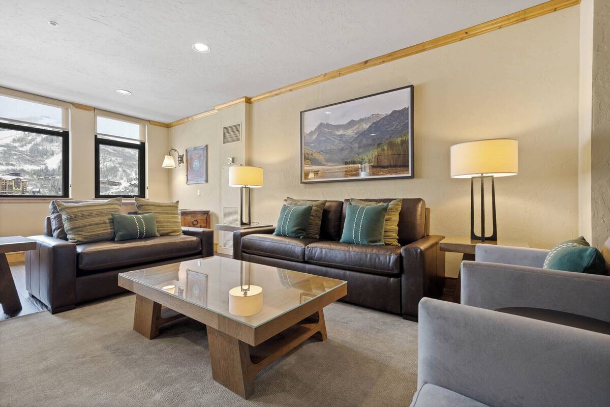 A modern living room with two leather sofas, a glass coffee table, and a view of mountains through large windows.