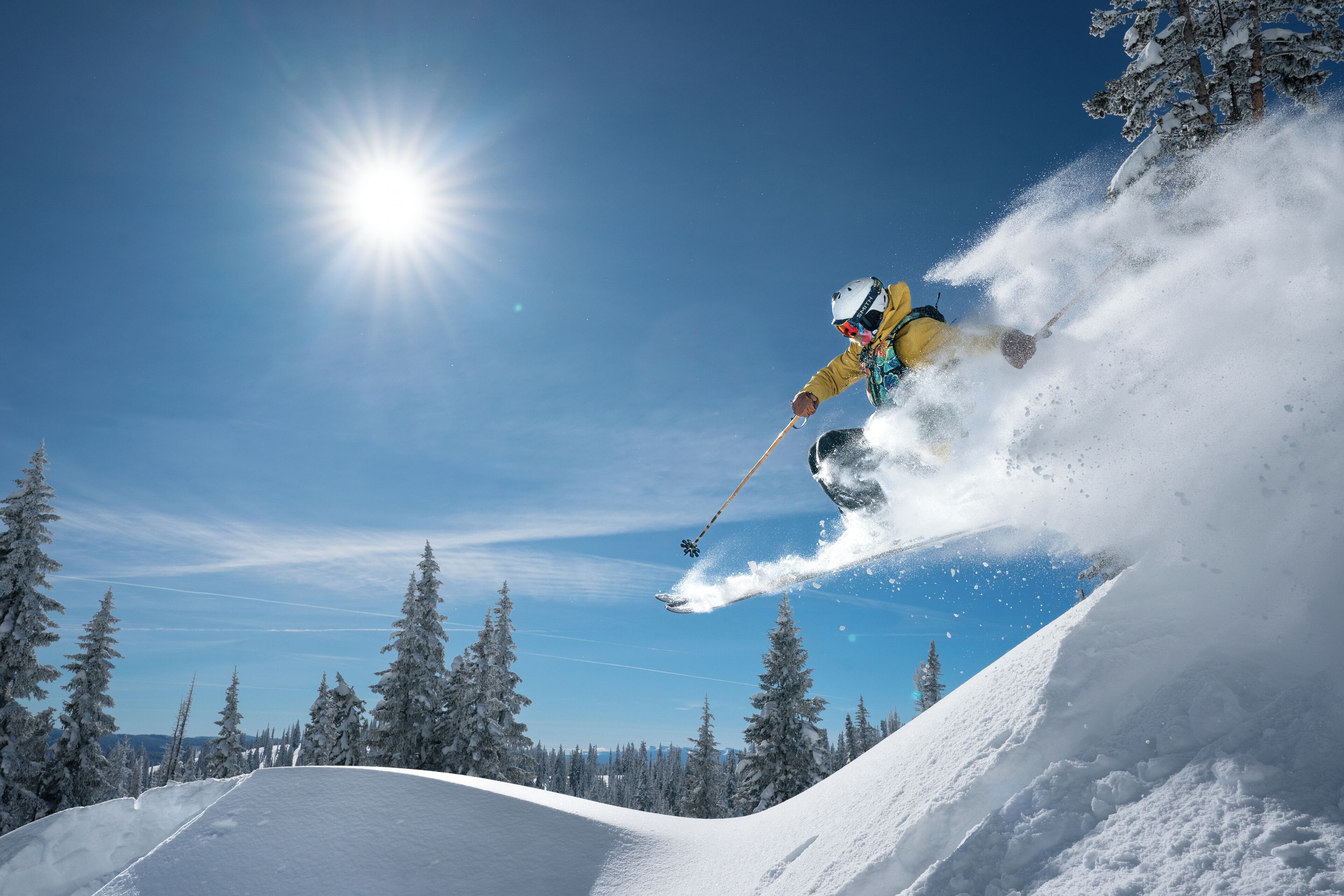 A skier in mid-air performing a jump on a snowy slope with trees in the background.