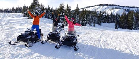 Tres personas en motos de nieve en una pendiente nevada, con árboles y una montaña al fondo.