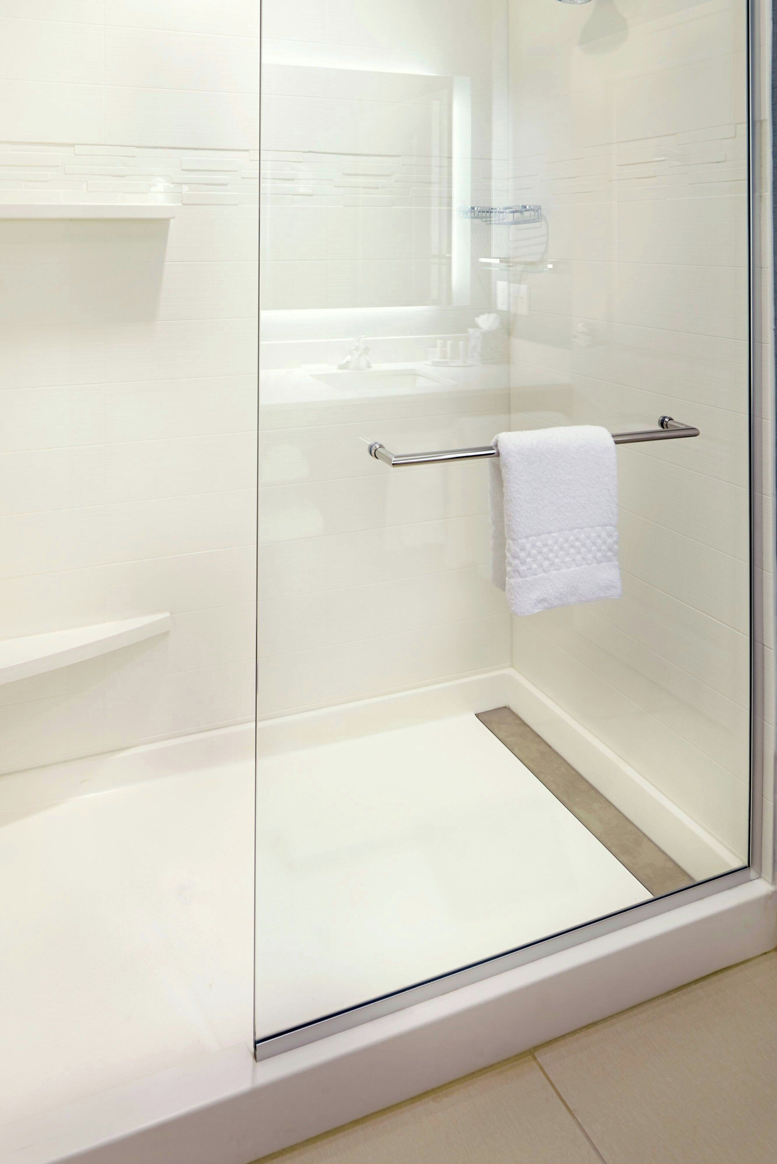 Ducha con mampara de vidrio, una toalla blanca sobre una barra de metal, un estante blanco y un recipiente de vidrio en la pared.