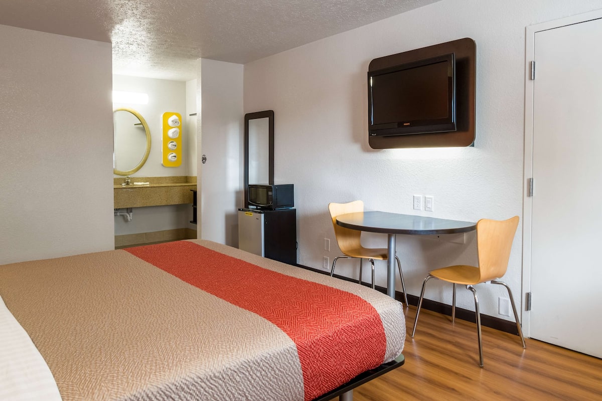 A hotel room with a bed, a desk, two chairs, a television, a mirror, and a small refrigerator.