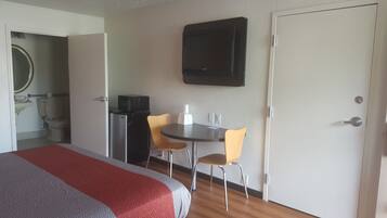 Habitación de hotel con cama, escritorio con dos sillas, microondas, refrigerador, televisor montado en la pared y una puerta.