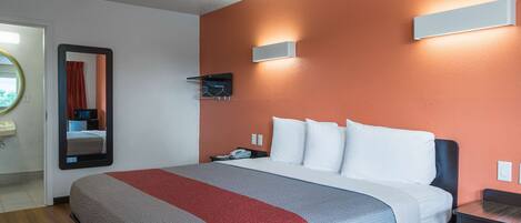 A hotel room with a bed, a bedside table, a mirror, and a wall-mounted light fixture.
