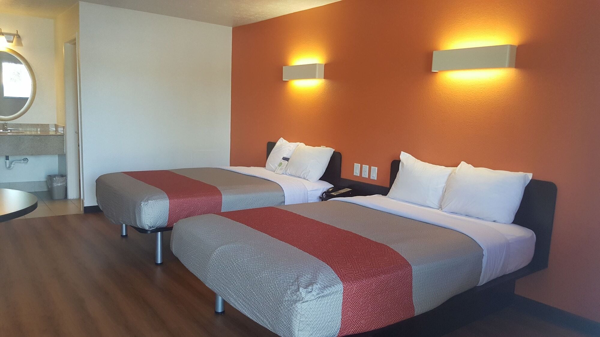 Habitación de hotel con dos camas, un espejo redondo, una mesita y una lámpara fijada a la pared.
