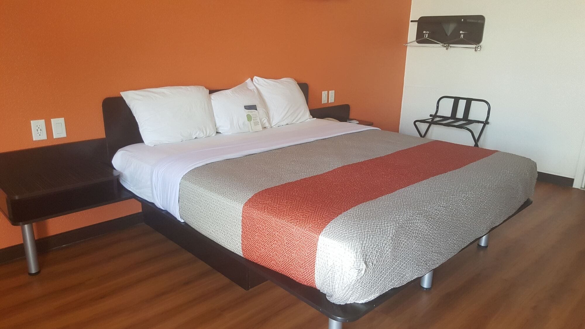 A bed with a white and gray blanket, a red and gray throw, and a black headboard. There is a small table with a chair beside the bed, and a wall-mounted TV above it.