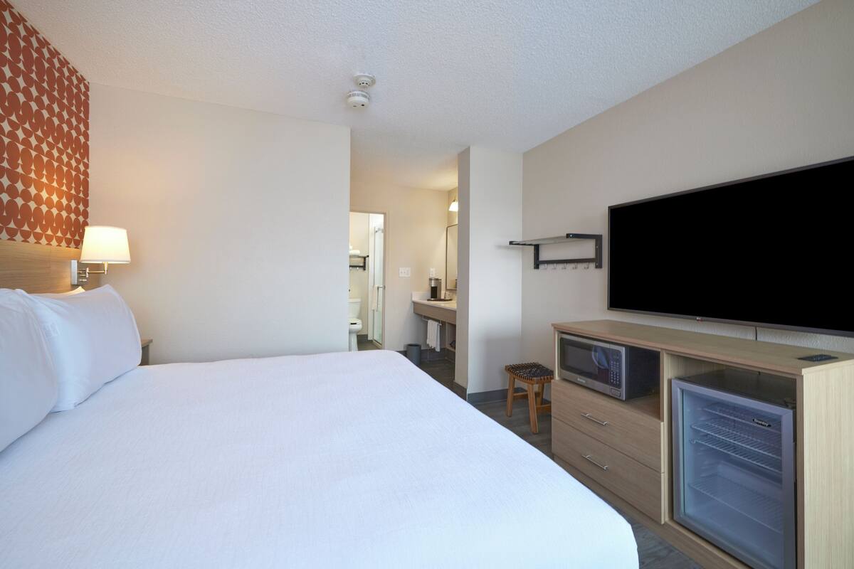 Une chambre d’hôtel avec un grand lit, un téléviseur à écran plat, un minibar, un petit tabouret et une salle de bain visible à travers une porte ouverte.