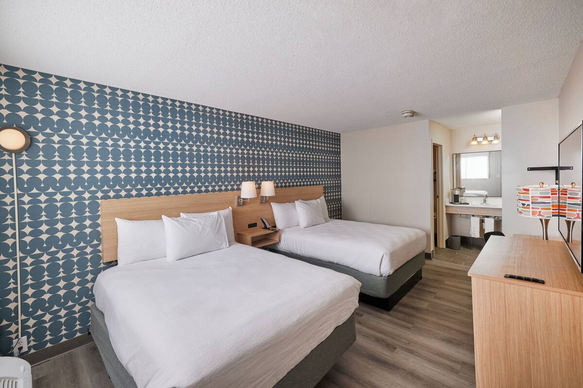 Une chambre d’hôtel avec deux lits, une tête de lit en bois, un mur à motifs, un bureau et une salle de bain visible à travers une porte ouverte.