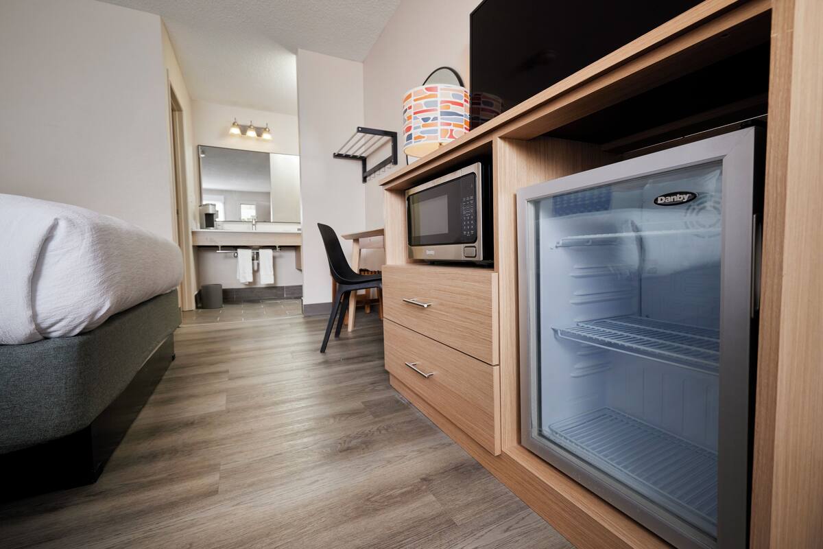 Une petite chambre comprenant un lit, un four à micro-ondes, un réfrigérateur, une chaise et une petite table avec une lampe.
