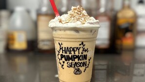 A pumpkin-themed beverage cup with a red straw, labeled "Happy Pumpkin Season," on a countertop with blurred bottles in the background. - Olentangy Motor Inn (Columbus)