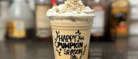 Un vaso con motivos de calabaza y pajita roja, con la etiqueta 'Happy Pumpkin Season', sobre una encimera con botellas desenfocadas al fondo.