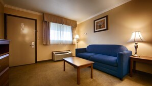 A hotel room with a blue sofa, a wooden coffee table, a standing lamp, a framed artwork on the wall, and a door with a number sign. - Best Western Clermont (Cincinnati)