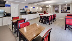 A modern hotel dining area with a long communal table and red cushioned chairs, a buffet station, and a television mounted on the wall. - Best Western Clermont (Cincinnati)
