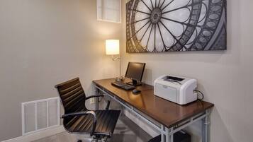 Habitación de hotel con escritorio, computadora, impresora, silla y lámpara de pared.