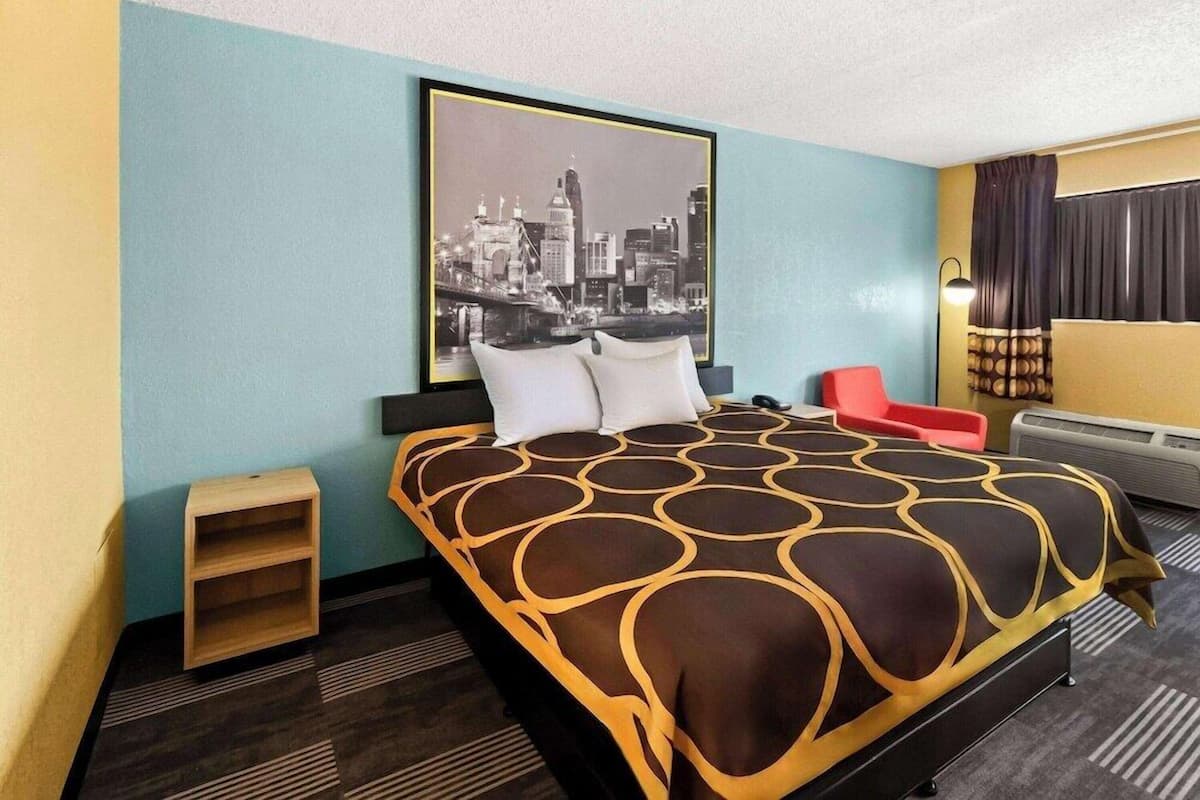 A hotel room with a bed, a cityscape picture on the wall, a small wooden shelf, and a red chair.