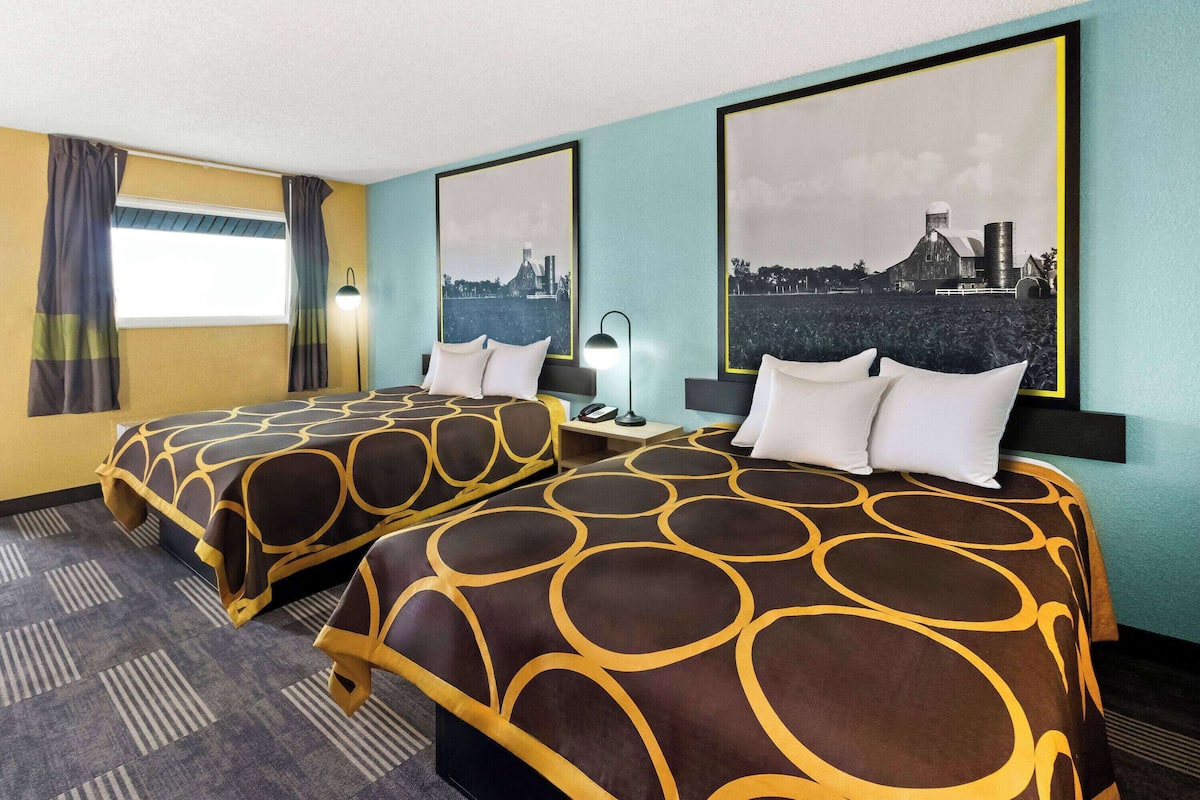 A hotel room with two beds, a large window with curtains, and two framed pictures on the wall.