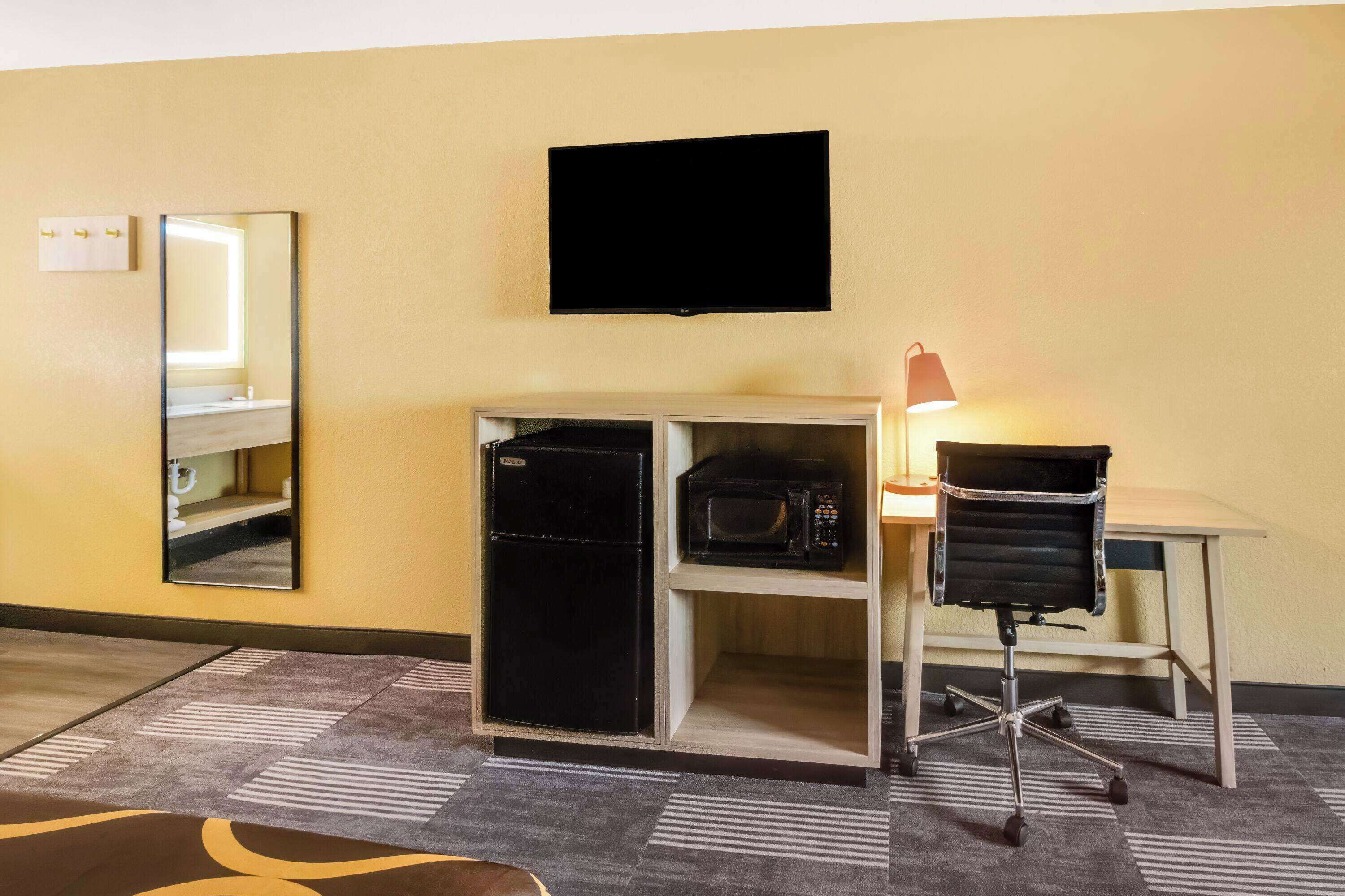 A compact hotel room with a kitchenette, a flat-screen TV, a mirror, and a desk with a chair.