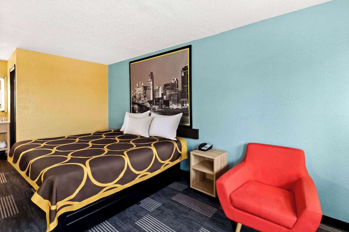 A hotel room with a bed, a red chair, a nightstand, and a cityscape painting on the wall.