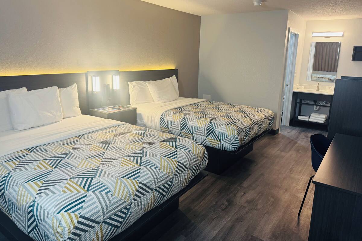 A hotel room with two beds, a desk, a chair, a bathroom with a sink and mirror, and a ceiling light.