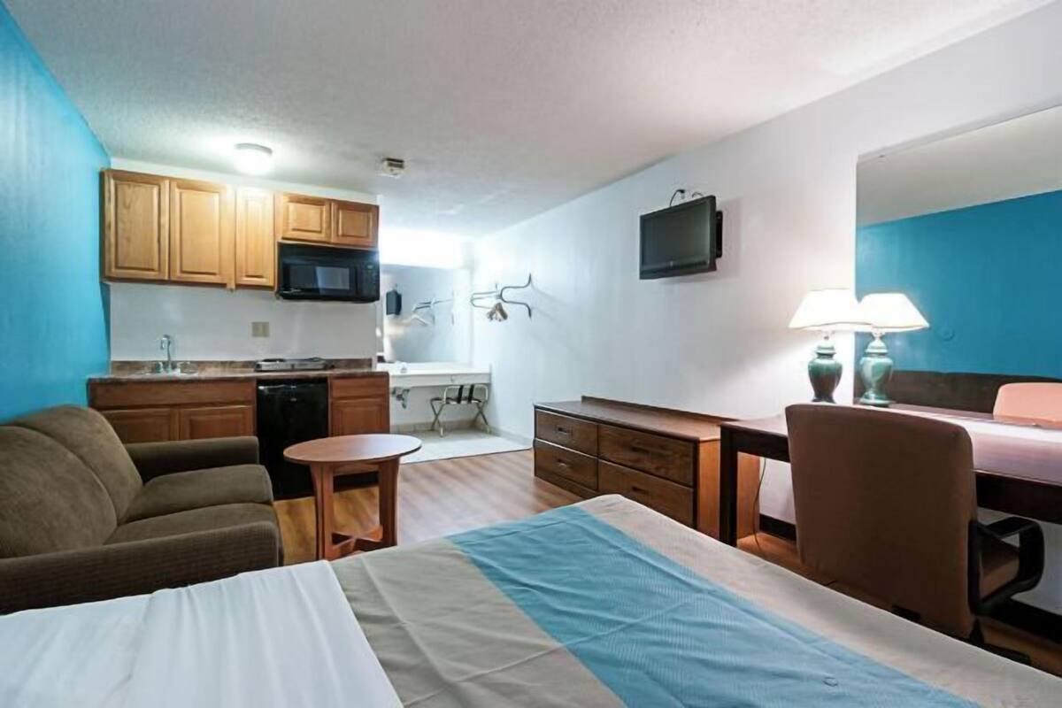A compact hotel room with a bed, a small desk, a chair, a sofa, a kitchenette, and a TV.