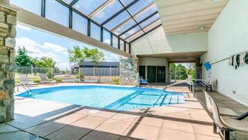 An indoor swimming pool with a glass ceiling, stone pillars, and a patio area with lounge chairs.