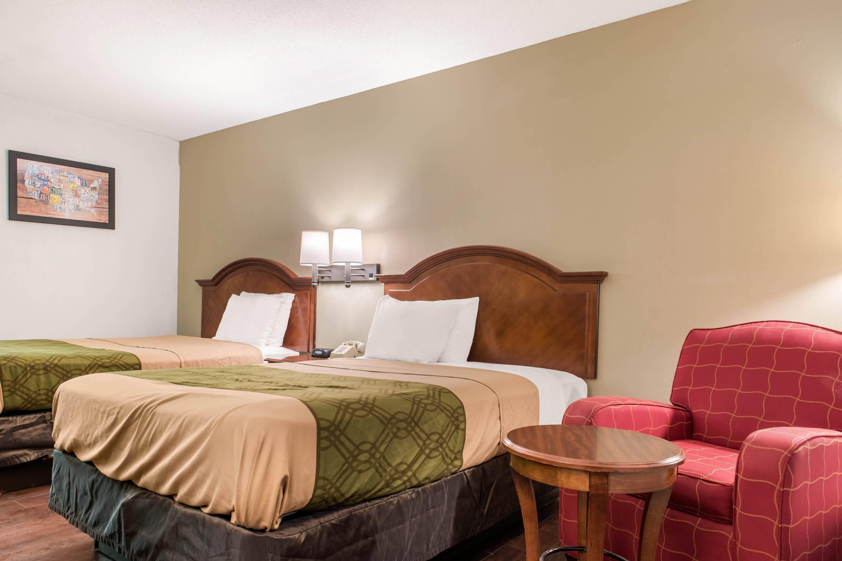 Une chambre d’hôtel avec deux lits, un fauteuil rouge recouvert de tissu, une petite table ronde et une lampe fixée au mur.