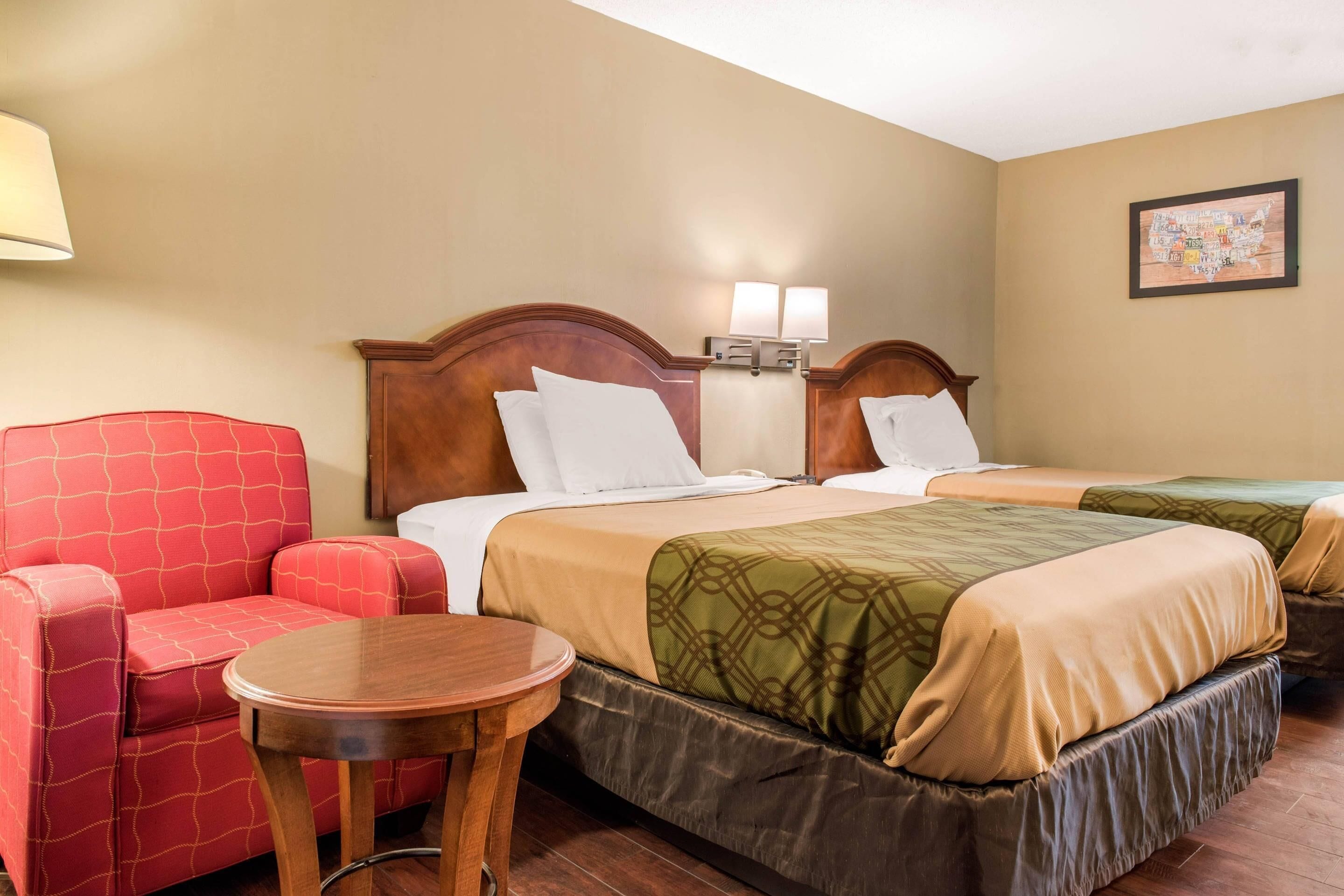 A hotel room with two beds, a red armchair, a small round table, and a wall-mounted lamp.