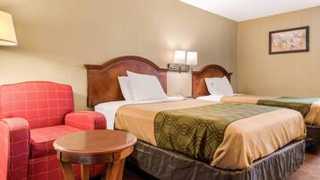 A hotel room with two beds, a red armchair, a small round table, and a wall-mounted lamp.